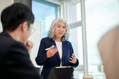 Profesyonel Asyalı kıdemli bir kadın CEO beyaz tahtanın önünde duruyor ve modern bir ofiste ekibine yeni bir iş kurma stratejisini özgüvenle anlatıyor..