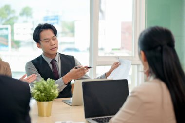 Resmi takım elbiseli profesyonel, zeki bir Asyalı işadamı, modern bir ofis ortamında yaptığı ortak bir iş toplantısında kendi fikirlerini paylaşıyor ve ekibine projesini anlatıyor..