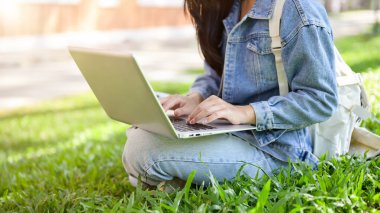 Üniversite parkındaki çimlere oturmuş dizüstü bilgisayarını kullanan, dizüstü bilgisayarında yazan kot ceketli bir üniversite öğrencisi görüntüsü. Üniversite hayatı, yaşam tarzları, eğitim