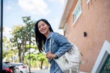 Çekici, gülümseyen, kot ceketli, sırt çantalı, sınıfına giden, açık havada üniversite alanında yürüyen Asyalı bir üniversite öğrencisi..