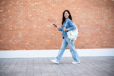 Kot pantolon giymiş güzel, gülümseyen Asyalı bir üniversite öğrencisinin yan görüntüsü. Sırt çantasını taşıyor ve dışarıda yürürken akıllı telefonunu tutuyor. Arka planda tuğla bir bina duvarı var..