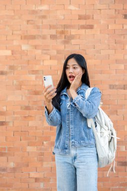 Şaşırmış, heyecanlı Asyalı bir üniversite öğrencisi arka planda tuğla bir duvarla dışarıda duruyor, akıllı telefon ekranına şaşırmış bir ifadeyle bakıyor, beklenmedik bir şekilde iyi haberler alıyor..