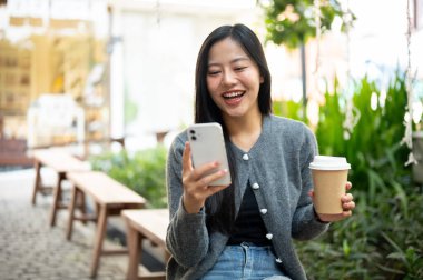 Hoş bir süveter giymiş çekici bir Asyalı kadın şehirdeki bir bankta oturmuş, akıllı telefonunda sohbet ederken sıcak bir içkinin tadını çıkarıyor. İnsanlar, yaşam tarzları, kablosuz teknoloji