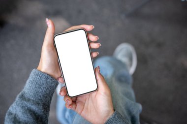 Üzerinde beyaz ekran modeli olan, arka planda bulanık bir caddesi olan akıllı bir telefonla dışarıda oturan, rahat gri kazaklı bir kadının üst görüntüsü. sohbet, uygulama, sosyal medya