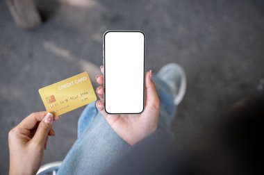 Açık havada oturan bir kadının üst görüntüsü, elinde kredi kartı ve arka planda bulanık bir sokak olan beyaz ekran maketli bir akıllı telefon. mobil bankacılık, online alışveriş, e-ticaret