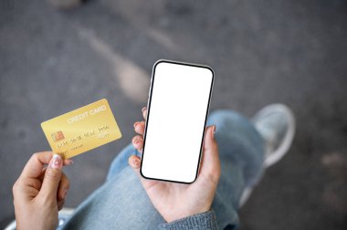 Açık havada oturan bir kadının üst görüntüsü, elinde kredi kartı ve arka planda bulanık bir sokak olan beyaz ekran maketli bir akıllı telefon. mobil bankacılık, online alışveriş, e-ticaret