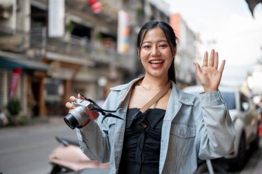 Çekici, kot ceketli mutlu bir Asyalı turist kamerasıyla fotoğraf çekerken şehri geziyor, gülümsüyor ve kameraya el sallıyor. insanlar ve yaşam tarzı kavramları