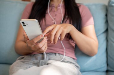 Parlak bir günde, bir kanepede oturan, akıllı telefonunu kullanan ve kulaklıklarıyla müzik dinleyen bir kadının yakın çekimi. İnsanlar, yaşam tarzları, teknoloji