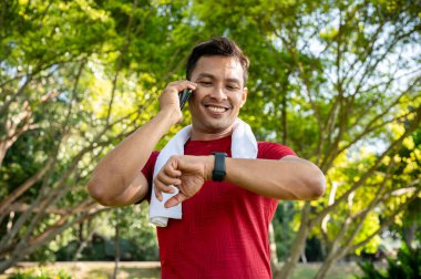 Spor kıyafetleri içinde sağlıklı, mutlu bir Asyalı adam telefonda konuşurken ve parlak bir günde parkta koşarken akıllı seğirmesini kalori sayısına göre kontrol eder. İnsanlar, yaşam tarzları, aygıtlar