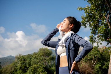 Spor kıyafetli yorgun, terli bir Asyalı kadın güneşli bir yaz gününde parkta egzersiz yaparken yorgun ve sıcak hissederek havluyla yüzünden ter siler. insanlar ve spor kavramları