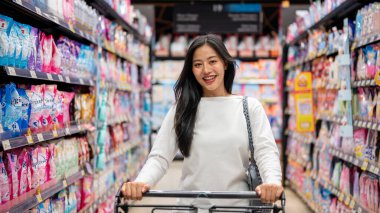 Chiang Mai, Tayland - 09 Aralık 2024: Çekici, gülümseyen Asyalı bir kadın süpermarketten alışveriş yapıyor, alışveriş arabasını itiyor, koridorda yürüyor ve hafta sonları ev eşyaları alıyor.