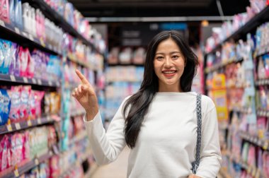 Chiang Mai, Tayland - 09 Aralık 2024: Güzel bir Asyalı kadın süpermarkette alışveriş yapıyor, alışveriş arabasıyla koridorda duruyor, kameraya gülümsüyor ve parmağını yukarı kaldırıyor..