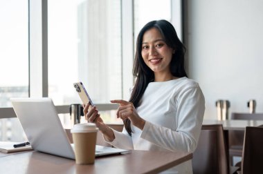 Modern bir binadaki bir kafede oturan çekici bir Asyalı kadın. Masasında dizüstü bilgisayarı ve elinde kameraya gülümseyen bir akıllı telefonla uzaktan çalışıyor. İnsanlar ve modern yaşam tarzı