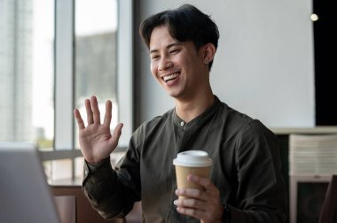 Neşeli bir Asyalı işadamı modern bir binadaki bir kafede çalışıyor ve ekibiyle çevrimiçi bir toplantı yapıyor. İnsanlar, modern yaşam tarzı ve kablosuz teknoloji kavramı