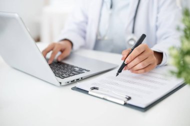 Ofisteki masasında çalışan, dizüstü bilgisayarının klavyesine yazı yazan bir kadın doktorun yakın plan fotoğrafı. Elinde eli masanın üzerindeki bir dosyada bulunan bir tıp kağıdını işaret eden bir kalem var..