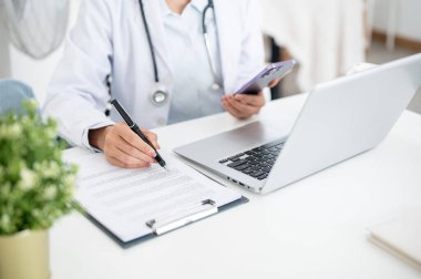 Ofisteki masasında çalışan bir doktorun kesilmiş bir görüntüsü. Akıllı telefonunu elinde tutarken, tıbbi belgeleri okurken, bilgisayarın başında otururken..