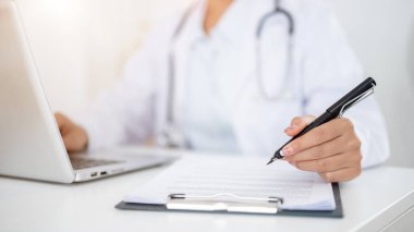 Ofisteki masasında çalışan, dizüstü bilgisayarının klavyesine yazı yazan bir kadın doktorun yakın plan fotoğrafı. Elinde eli masanın üzerindeki bir dosyada bulunan bir tıp kağıdını işaret eden bir kalem var..