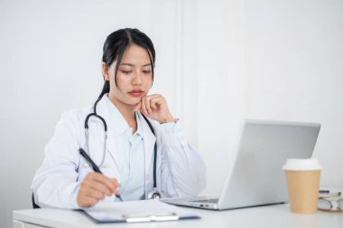 Üniformalı çekici bir Asyalı kadın doktor, tıbbi vakaları kağıt ve dizüstü bilgisayarında okumaya odaklanmış, hastanedeki ofisinde çalışıyor. sağlık hizmeti kavramı
