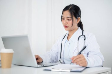 Üniformalı çekici bir Asyalı kadın doktor, tıbbi vakaları kağıt ve dizüstü bilgisayarında okumaya odaklanmış, hastanedeki ofisinde çalışıyor. sağlık hizmeti kavramı