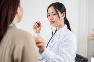 Profesyonel bir Asyalı kadın doktor, hastanın akciğer ve kalp atışlarını dinlemek için steteskop kullanıyor. Hastanedeki bir muayene odasında, onları hastalık belirtileri için dikkatlice inceliyor..