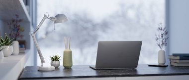 Bir dizüstü bilgisayar, bir masa lambası ve modern bir ofis çalışma alanındaki bir masanın üzerine karlı bir kış manzarası manzarası sunan geniş bir pencereyle bir dekor. 3d görüntüleme, 3d illüstrasyon
