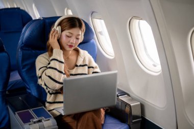 Cam kenarında oturan, kulaklık takan ve uçuş sırasında dizüstü bilgisayarı üzerinde çalışan güzel Asyalı bir kadın..