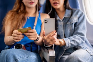 Uçakla seyahat eden iki Asyalı kadının yakın plan fotoğrafı. Biri elinde telefon, diğeri de kredi kartı tutuyormuş. Uçuş sırasında kredi kartının kolaylığı hakkında konuşuyorlarmış..