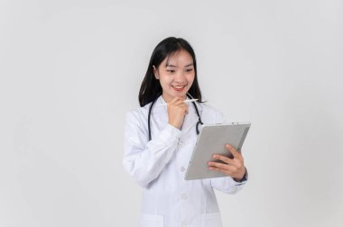 Beyaz önlüklü güzel, pozitif Asyalı bir kadın doktor beyaz bir stüdyonun arka planında duruyor, dijital tabletinde tıbbi vakaları okuyor..