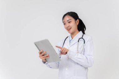 Beyaz önlüklü, güzel, gülümseyen bir Asyalı kadın doktor dijital tabletini kullanıyor, tıbbi vakaları okuyor, beyaz bir stüdyonun arka planında duruyor..