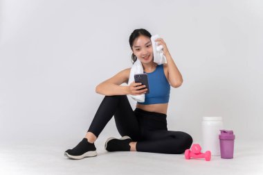 Spor kıyafetleri içinde güzel, sağlıklı bir Asyalı kadın beyaz bir stüdyoda oturuyor, akıllı telefonunu kullanırken yüzünden terini siliyor, idmandan sonra dinleniyor..