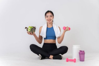 Spor kıyafetleri içinde mutlu, sağlıklı bir Asyalı kadın bir elinde bir dambıl diğer elinde de yanında protein içeceği olan bir salata kasesi olan beyaz bir stüdyoda oturuyor. sağlıklı yaşam biçimleri