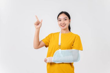 Kırık koluyla gülümseyen Asyalı bir kadın, yara bandına sarılı ve bir kazadan sonra boş bir yeri işaret eden bir askıyla destekleniyor. Stüdyo geçmişi, sağlık hizmeti kavramı