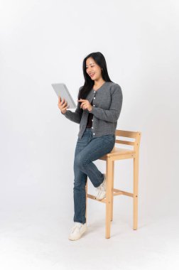 Rahat giysiler içinde çekici bir Asyalı kadın elinde dijital tabletle beyaz bir arka planda tahta bir sandalyede oturuyor. Film izlemek, uzaktan çalışmak.