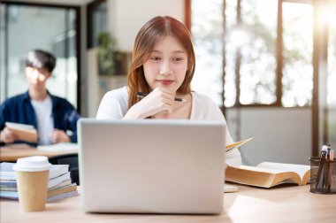 Gülümseyen, mutlu Asyalı bir üniversite öğrencisi bilgisayarında, bir kahve dükkanında ya da kütüphanede internetten ders çalışıyor. insanlar ve eğitim kavramları