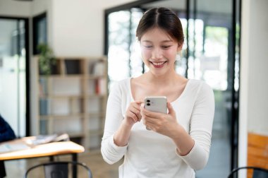 Güzel, gülümseyen Asyalı genç bir üniversite öğrencisinin portresi. Günlük kıyafetlerle kütüphanede dururken akıllı telefonundan mesajlar okuyor. insanlar ve eğitim kavramları