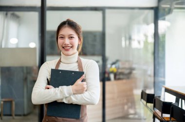 Çekici bir Asyalı kadın kafe çalışanı ya da rahat kıyafetler giyen küçük bir işletme sahibi kahve dükkanında kameraya gülümsüyor..