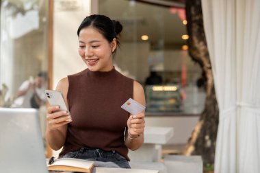 Mutlu, tatmin olmuş Asyalı bir kadın bir kafede otururken akıllı telefonuyla alışveriş yapmaktan hoşlanır. Masanın üstünde bir kredi kartı ve akıllı telefonu vardır. Çevrimiçi ödeme, e-ticaret, yaşam tarzı, mali
