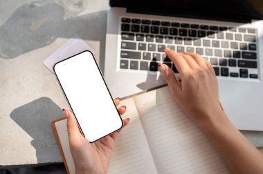 Üzerinde beyaz ekran modeli olan akıllı telefonu elinde tutan bir kadının üst görüntüsü. Üzerinde bir not defteri ve dizüstü bilgisayarı olan, çalışırken akıllı telefonunu kullanan..