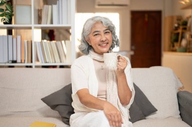 Güzel, gülümseyen Asyalı son sınıf öğrencisi, rahat giysiler içinde, elinde kahve fincanıyla oturma odasındaki kanepeye oturmuş kameraya gülümsüyor. İyi yaşam, mutlu emeklilik, ev hayatı.