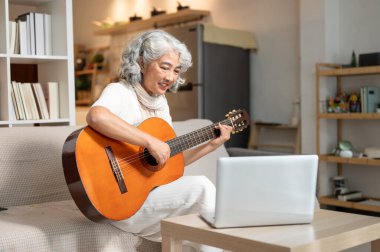 Olumlu, rahat Asyalı son sınıf öğrencisi bir kadın evde dizüstü bilgisayarından ders çalışırken oturma odasında akustik gitar çalışıyor. Boş zaman, yaşam tarzları, emeklilik