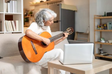Olumlu, rahat Asyalı son sınıf öğrencisi bir kadın evde dizüstü bilgisayarından ders çalışırken oturma odasında akustik gitar çalışıyor. Boş zaman, yaşam tarzları, emeklilik