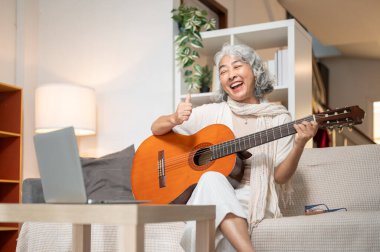 Neşeli bir Asyalı emekli kadın evdeki bilgisayarında akustik gitar dersi alıyor oturma odasında bir kanepede oturuyor ve gitar çalıyor..
