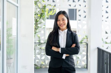 Resmi bir iş elbisesi içindeki kendine güvenen, başarılı Asyalı bir iş kadınının portresi koridorda kollarını kavuşturup kameraya gülümsüyor..