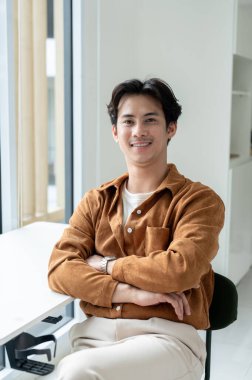 Yakışıklı, pozitif Asyalı bir adam kollarını kavuşturup bir odada kameraya gülümseyen bir masada oturuyor. Ofiste bir işadamı var..