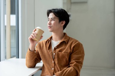 Yakışıklı, düşünceli Asyalı bir adam minimalist bir kafede kahvesini yudumlarken pencereden dışarı bakıyor. insanlar ve yaşam tarzları kavramları