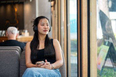 Güzel, rahat bir Asyalı kadın bir kahve dükkanında kulaklıklarıyla müzik dinlerken pencerenin kenarında oturur. insanlar ve yaşam tarzları kavramları