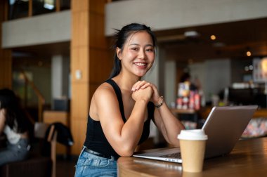 Çekici bir Asyalı kadın kahve dükkanında uzaktan çalışıyor, bir masada dizüstü bilgisayarıyla oturuyor ve kameraya gülümsüyor. İnsanlar, yaşam tarzı, kablosuz teknoloji