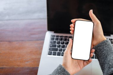 Bir kadının yakın çekimi, ahşap bir masanın üzerindeki dizüstü bilgisayarın üzerinde beyaz ekran modeli olan bir akıllı telefonu tutuyor. İnsanlar, yaşam tarzları, kablosuz teknoloji