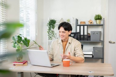 Olumlu, kendine güvenen Asyalı bir işadamı ofisinde çalışır ve müşterisiyle bilgisayarı aracılığıyla çevrimiçi bir toplantı yapar. Uzaktan çalışan kablosuz teknoloji.