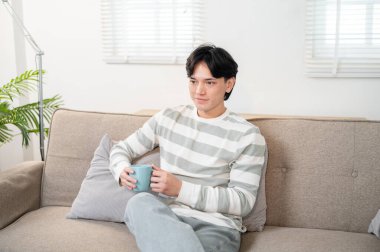 Yakışıklı, mutlu Asyalı genç bir adam TV 'de haberleri izlerken oturma odasındaki kanepede sabah kahvesinin tadını çıkarıyor. ev hayatı, yaşam tarzı